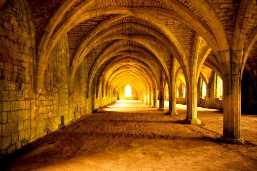 Bright golden light shines through the arched window at the end of the Cellarium at Fountains Abbey, North Yorkshire, England. Fountains Abbey was founded in 1132 following a dispute and riot at St Mary's Abbey in York. Following the riot, thirteen monks were exiled they founded Fountains Abbey which operated for over 400 years, until 1539, when Henry VIII ordered the Dissolution of the Monasteries.
