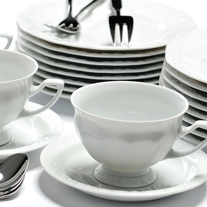 Close-up of very elegant china tableware (plates, dessert plates, cups, spoons and forks) on white table. Studio shot, black and white.