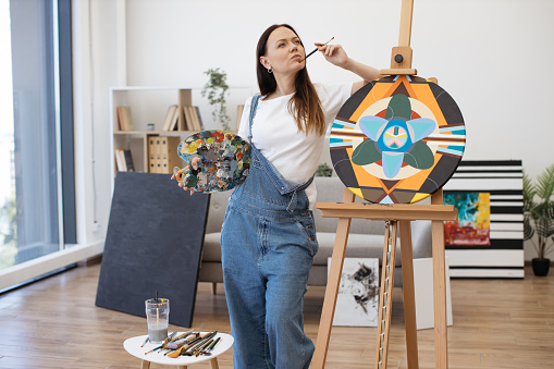 Pensive caucasian woman touching chin with paintbrush and leaning with elbow on wooden easel indoors. Pretty female holding colored palette and thinking about ideas for new artworks.