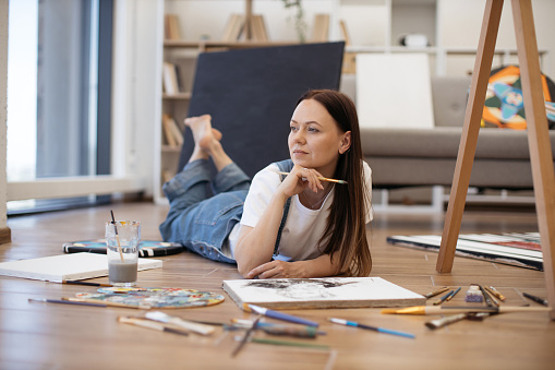 Beautiful woman with long brown hair lying on stomach with crossed legs and looking at window. Creative painter in casual attire holding paintbrush in hand and looking for inspiration.