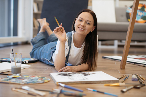 Concentrated caucasian lady lying on floor with painting accessories and drawing indoors. Dark-haired female artist dedicating weekend time for hobby. Concept of creativity and inspiration.