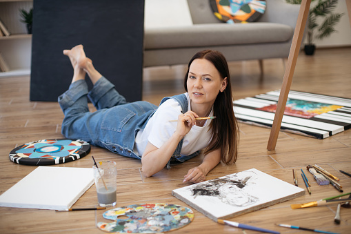 Young attractive woman dressed in white t-shirt and denim jumpsuit lying on floor of cozy apartment and posing on camera with paintbrush. Talented artist drawing picture while staying at home.