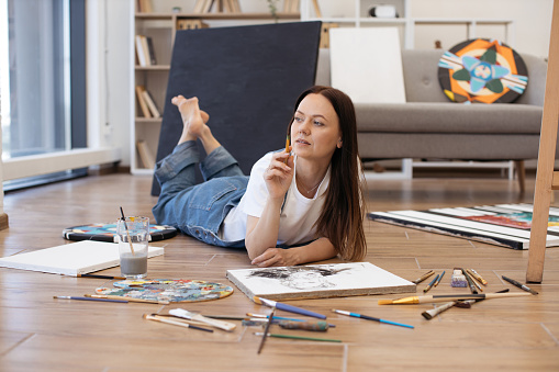 Beautiful woman with long brown hair lying on stomach with crossed legs and looking at window. Creative painter in casual attire holding paintbrush in hand and looking for inspiration.