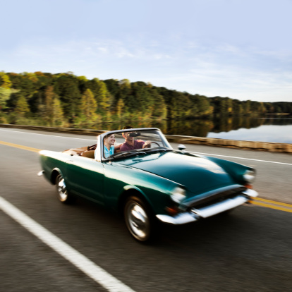 Couple taking a road trip in a vintage convertible car and driving pass a scenic view of trees and water. Square shot.