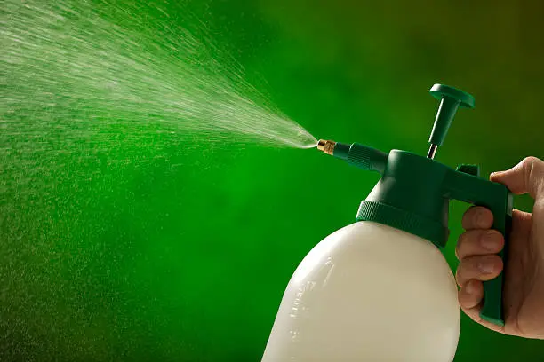 A man is holding a plastic garden spray bottle and spraying water out of the nozzle. Horizontal shot.