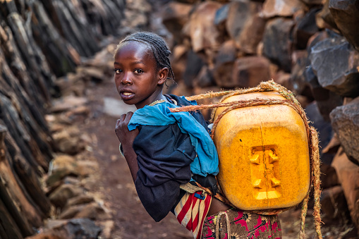 African girl from Konso people carrying water to the village, African women and children often walk long distances to bring back jugs of water that they carry on their back. 
She lives in a stone village which is a part of The Konso Cultural Landscape in the Konso highlands of Ethiopia (UNESCO Heritage). Each of these villages has stone walled terraces and fortified houses.