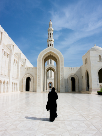 Woman in black Abaya Gown walking towards the giant arch in the famous Sultan Qaboos Grand Mosque in Muscat, Oman, Middle East.
