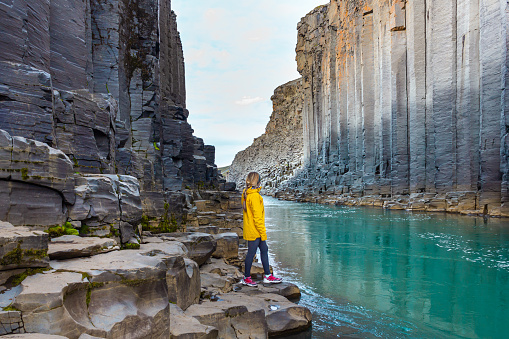 Hiker woman in yellow jacket visiting Studlagil Canyon, Iceland. Famous touristic landmark destination with volcanic basalt columns.