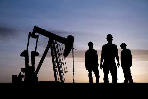 Three men who work in the oil and gas field have a meeting as a pumpjack works behind them.