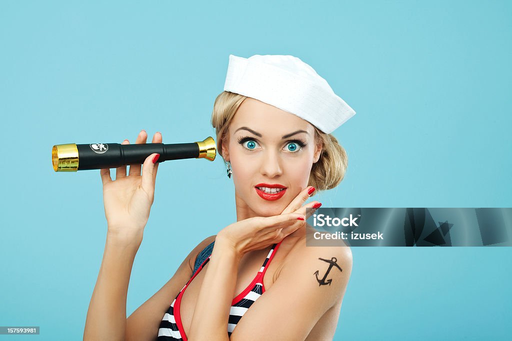 Pin-up style sailor woman holding telescope Young Blond Woman with anchor tatoo on her arms Wearing Striped Bikini and Blue Overalls holding telescope in hand. Standing against blue background. Pin-Up style. Summer portrait. Sailor Stock Photo