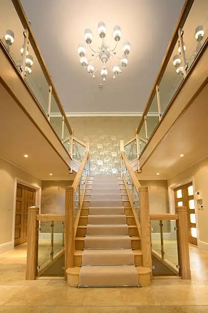 a grand modern staircase in an expensive new home. Built from solid oak with glass panels and chromed fittings, this is a contemporary style for luxury new houses. A modern light cluster on the ceiling defers to the traditional chandelier. The floor area comprises large flagstones which add to the yellow feel to the scene.
