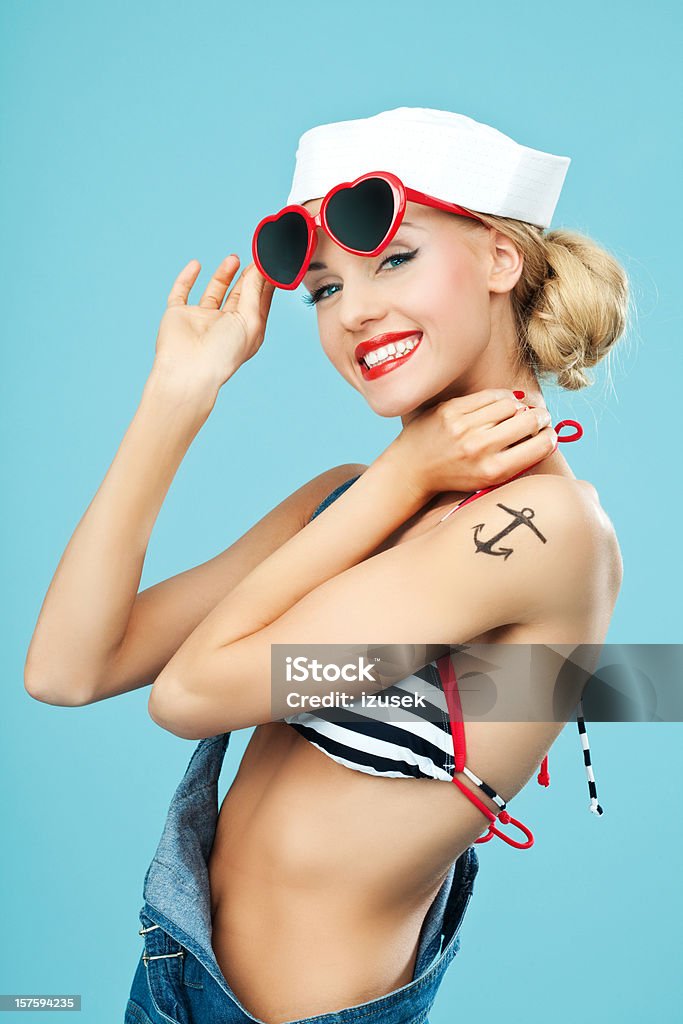 Pin-up style sailor woman with sunglasses Young Blond Woman with anchor tatoo on her arms Wearing Striped Bikini, Blue Overalls and sunglasses. Standing against blue background. Pin-Up style. Summer portrait. Sailor Stock Photo