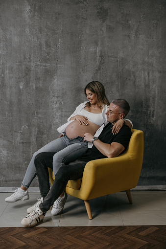 Full length shot of happy pregnant couple sitting on chair and relaxing at home