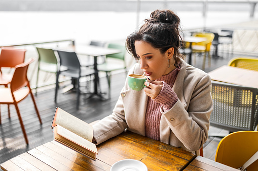 Young woman sitting in cafe drinking coffee reading book
