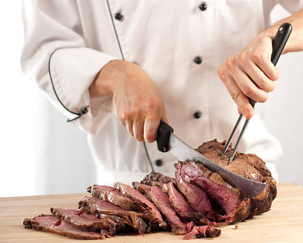 Chef slicing roast beef using carving knife and fork Professional chef carving slices of a perfectly cooked medium rare prime rib roast beef using a carving knife and fork in a commercial kitchen that could be at a restaurant, hotel, cooking school, café or catering operation in the food and beverage industry. carving food photos stock pictures, royalty-free photos & images