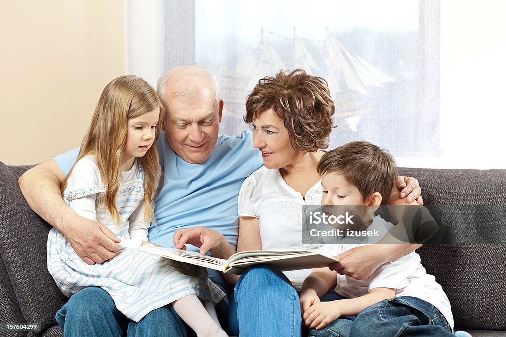 Grandparents with grandchildren reading book Grandparents with grandson and granddaughter sitting on sofa in living room and reading book. Children listening with great interest, grandparents smiling. 6-7 Years Stock Photo