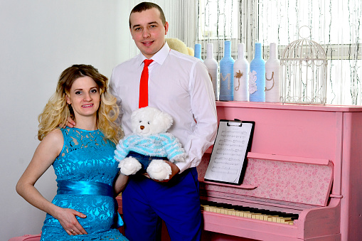 Attractive young couple is celebrating holiday at home. Standing on the background of beautiful a pink piano. Husband hugs his pregnant wife. A man and his pregnant wife stand by a piano. Standing and smiling near a pink piano. High quality photo.