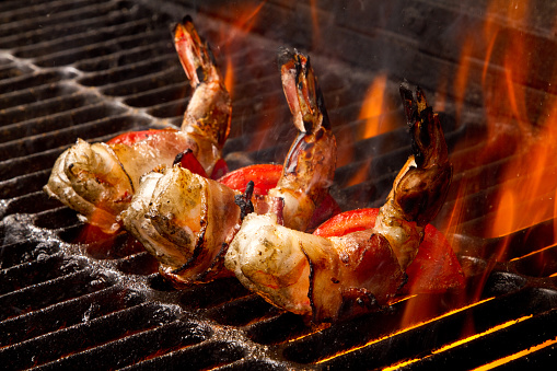 Row of three succulent jumbo shrimp or prawns wrapped in bacon on an old fashioned charcoal grill with flames leaping up to sear them.  Cooking is bringing out a golden brown and red color on the shrimp.