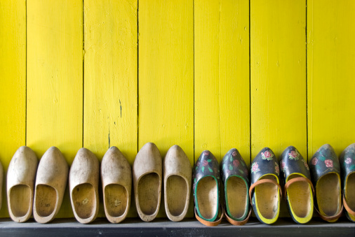Row of pairs of used wooden shoes placed in line against a bright yellow wooden wall. Some wooden shoes are painted in traditional dutch pattern. Others are blank. The wooden shoes are part of the dutch culture in the Netherlands.