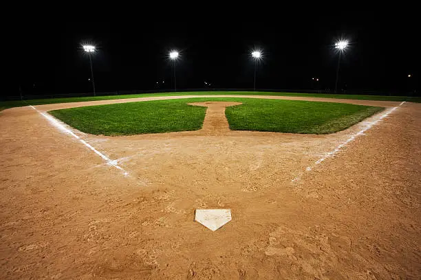 Groomed softball and baseball diamond at night.  Bright stadium lights illuminate the red sand and green grass field while darkness isolates the field from its surroundings.  No players on the field.