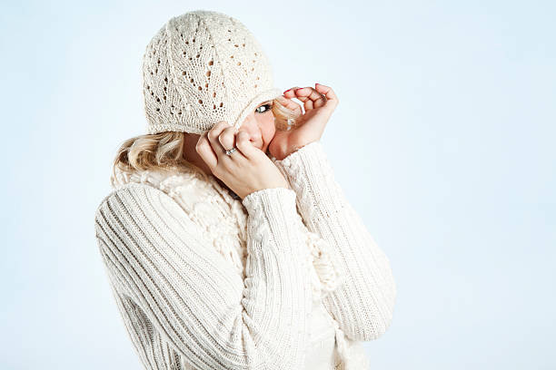 Photo of a young woman in soft sweater, knit cap and scarf, posing against a bright blue background; peeking from behind cap pulled over her eyes.