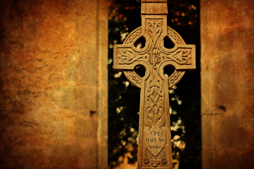 A cross on a weathered gravestone in a rural churchyard in bright sunlight. The gravestone is covered in lichen and moss which almost obscures the carving.