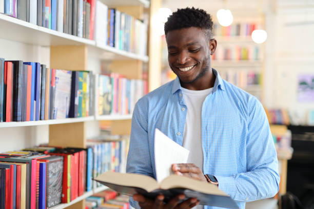 istock Black man in a library or bookstore holding an open book and reading it while wearing a blue button-down shirt. The background consists of bookshelves filled with colorful books. 1576109887