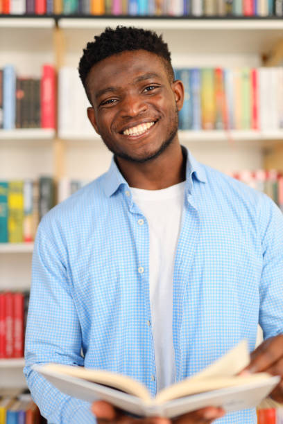 istock Black student in a library holding an open book while wearing a blue collared shirt. The background consists of bookshelves filled with colorful books. 1576110059