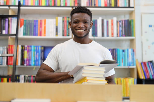istock Smiling black guy with books in the library. 1576111522