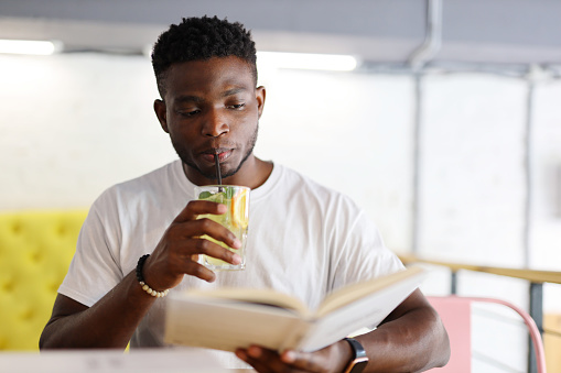 African man sitting in a cafe and reading a book while holding a glass of drink. He is a young and happy student, businessman or ethnic who is studying, working or learning for his education.