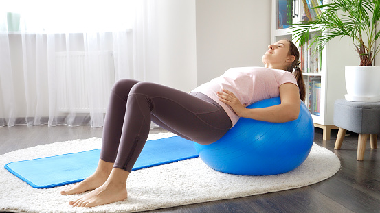 Young woman in leggings stretching back muscles while rolling on blue fitness ball.