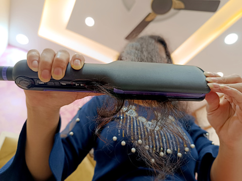 Young women of Indian ethnicity straightening hair with using hair Straightener at home.