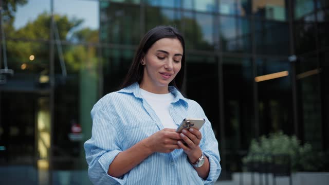 Beautiful young business woman walking texting typing message on mobile phone outside business center. Female tapping scrolling on smartphone and stepping. Businesswoman using app for social network.