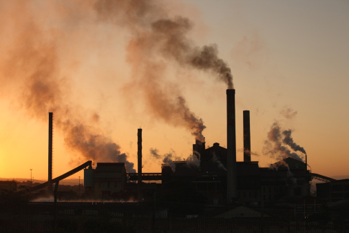 Backlit paper mill at sunset with dark smoke rising upwards into the orange sky