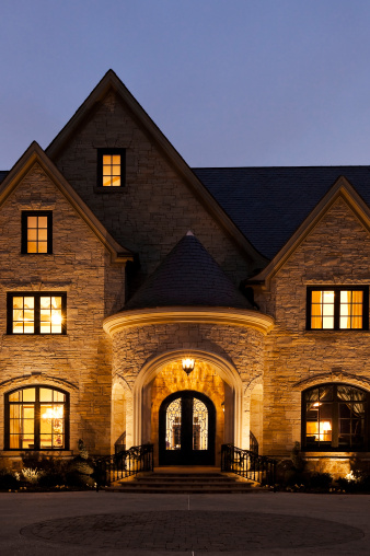The front door of a large mansion with a circular driveway at night.  Exterior and interior lights are on. Vertical shot.