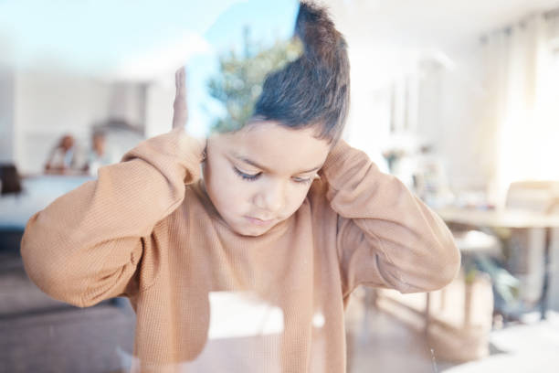 Sad, home and boy with hands on ears for depression, fear and scared of conflict with family by window. Child psychology, autism and depressed, anxiety and upset kid in living room for mental health stock photo