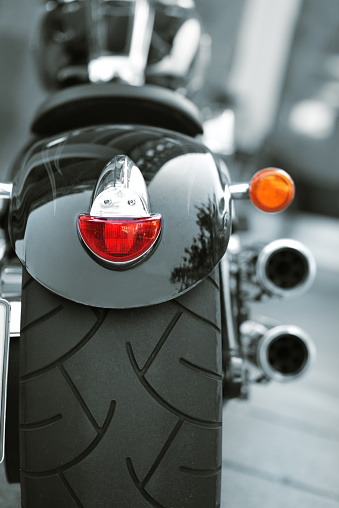 Rear view from of a very large motorcycle tire from an parked black motorcycle. This type of tire are typical for harley davidson motorcycles. Vertical shot with focus on tire. Álso you can see the red rearlight, the exhaust pipes and a orange blinker on the right side. Shooted outdoors in daylight with full format camera and prime lens. Processed in raw. Shallow depth of field 