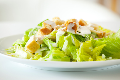 Detail of a classic chicken caesar salad with lettuce, chicken, croutons, caesar dressing and a sprinkle of pepper in kitchen setting.