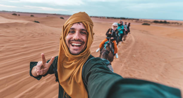 turista felice che si diverte godendo di un giro in cammello di gruppo nel deserto - viaggi, stile di vita, attività di vacanza e concetto di avventura - morocco desert camel africa foto e immagini stock