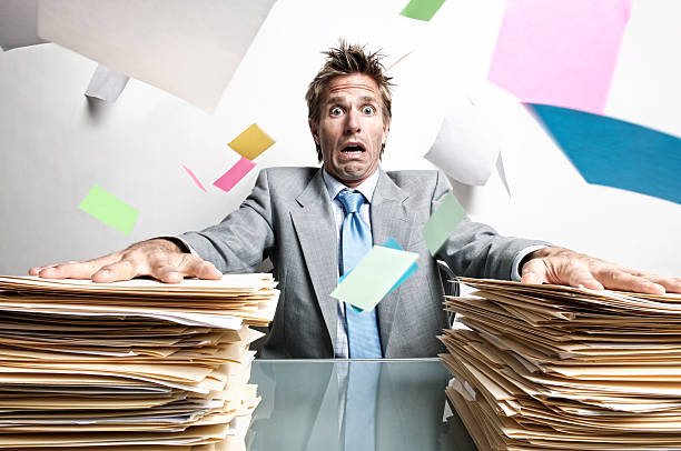 Stressed businessman office worker panicking trying to keep his papers down as his inbox flies into the air around him  in chaos on his desk
