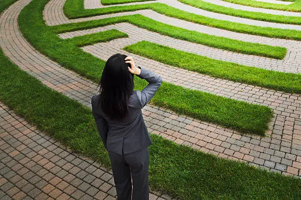 A business woman stands scratching her head, wondering in front of a grass maze, perhaps lost or searching for a solution to her business problem. Illustrates concepts for business problem-solving and management strategy, or employment and occupation issues, uncertainties, and problems.