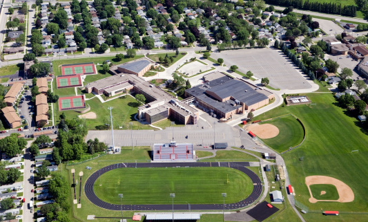 This is an aerial shot of a large public high school complex shot on a Sunday with nobody around. This image features multiple buildings, a running track, football fields, baseball diamonds, tennis courts parking lots and a residential neighborhood surrounding the image. Shot from the open window of a small plane.