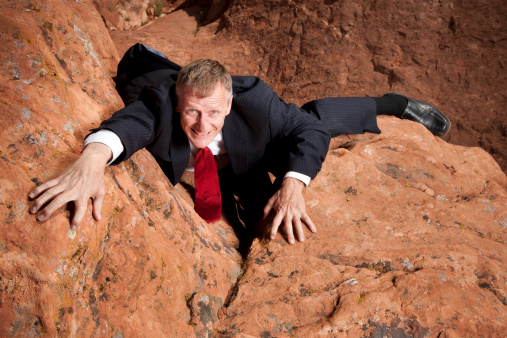 High angle view of a businessman in a suit and tie struggling to climb up a steep cliff.