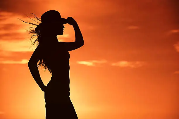 Cowgirl Silhouette in a hot orange Western Sky. A bold back light was created around her by placing the sun directly behind the woman.  The wind is blowing her hair, and this background has plenty of copy space in a moody sunset. 