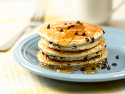 Pancakes with syrup and chocolate chips on blue plate.  Coffee cup and fork in background.  Selective focus.