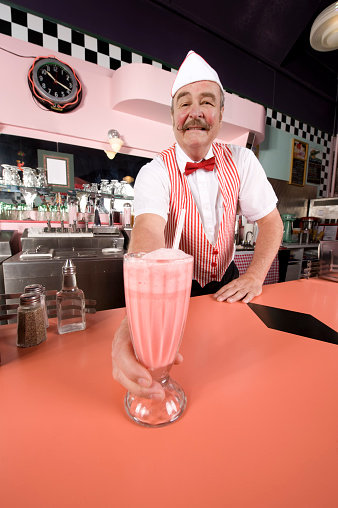 Photograph in a 1950, 1960s era soda shop with a waiter serving you a milkshake. Shades of pink accented with a strawberry milkshake.  Older man with a red and white stripped vest and red bow tie serving you.  Copy space.