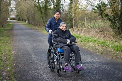 Asian Indian woman pushing her elderly mother in a wheelchair outdoors in winter, UK. May also depict a carer, care in the community