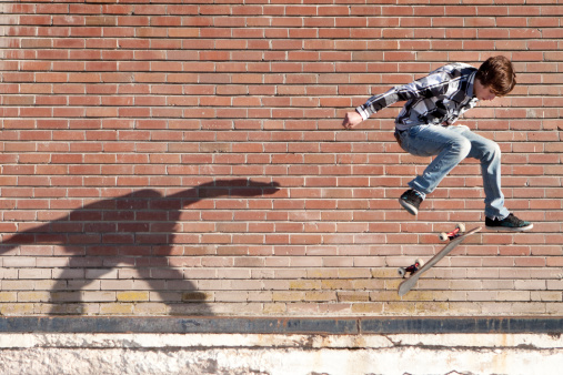 Male teenager doing a jump on skateboard against red brick wall, with his shadow on background.