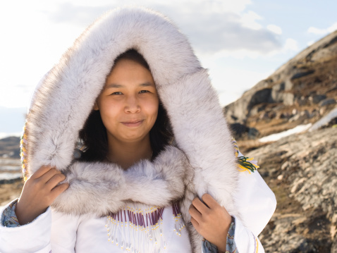 Picture of an Inuit woman standing on a tundra in Baffin Island, Canada.  The woman is wearing a thick fur hood over thick white traditional clothing.  The woman is wearing a necklace decorated in white and purple.  The sky above the tundra is filled with white clouds.