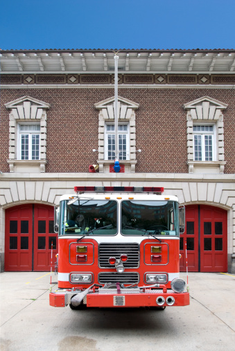 Front view of a red fire truck parked symmetrical in front of a fire station. With a clear blue sky.